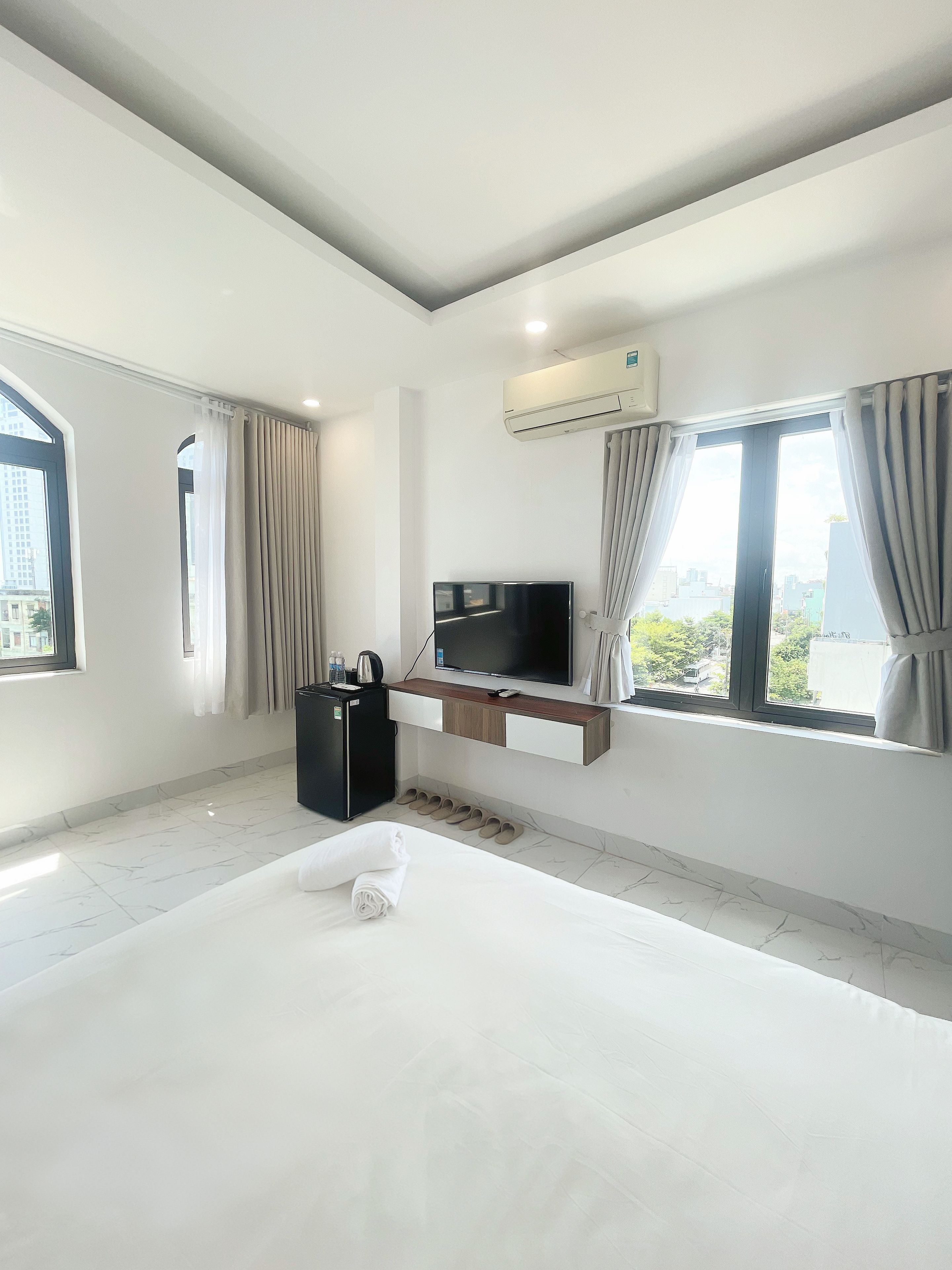 A modern hotel room with a bed, a flat-screen TV, a minibar, and a view of the city through large windows.