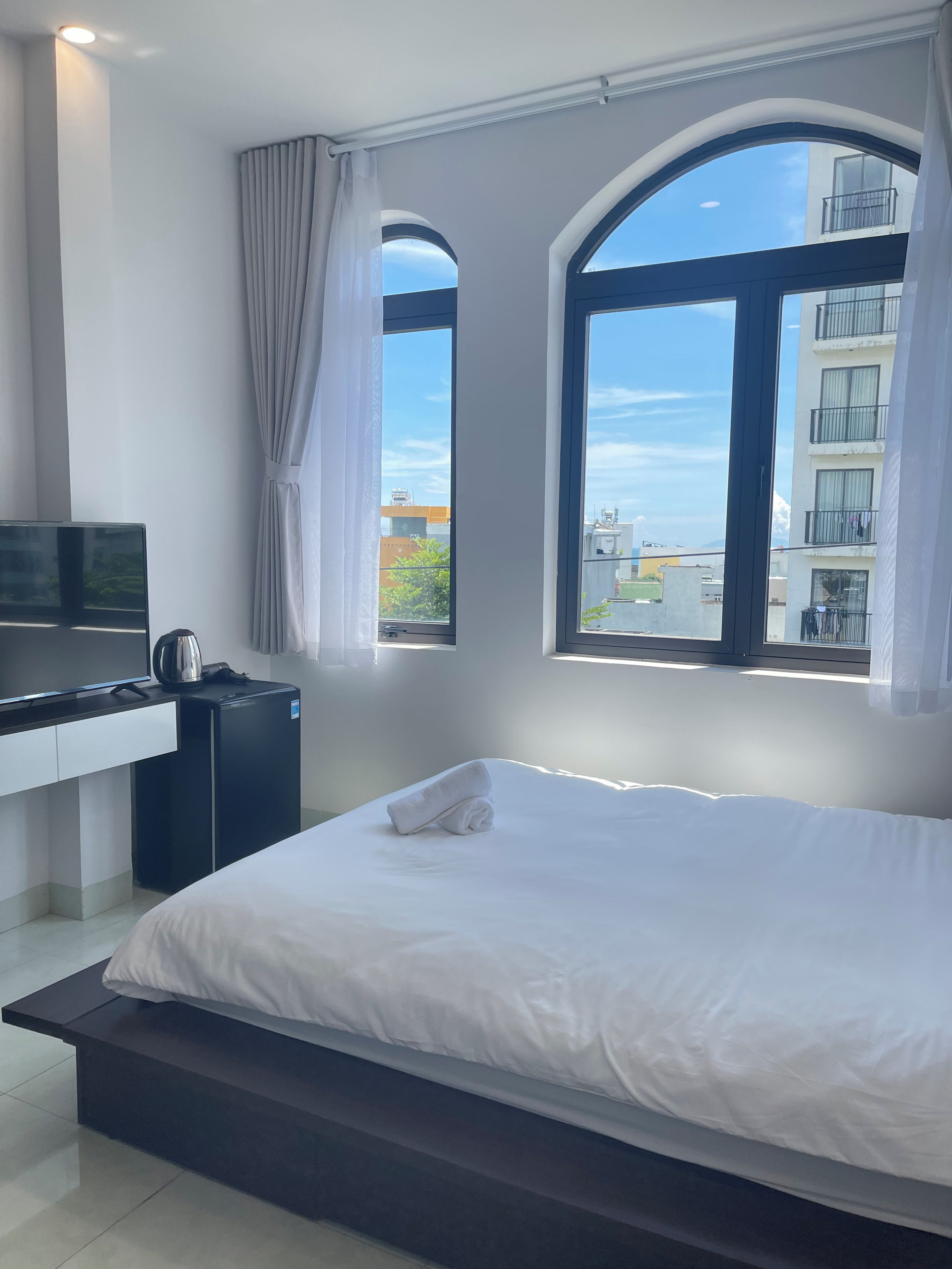 A hotel room with a bed, a desk, a television, and a view of buildings through large windows.