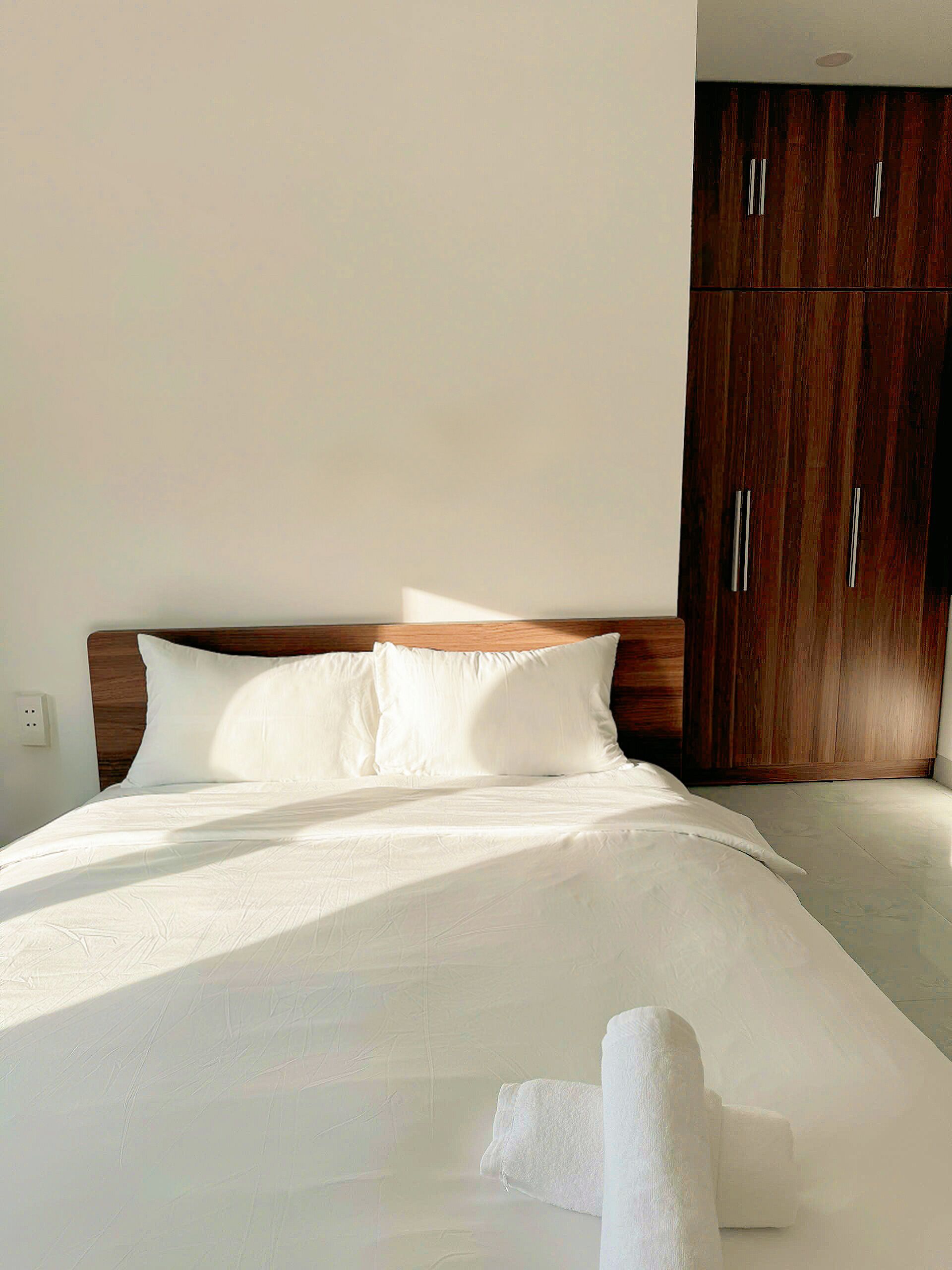 A neatly made bed with white linens and pillows, a wooden headboard, and a large wardrobe with silver handles.