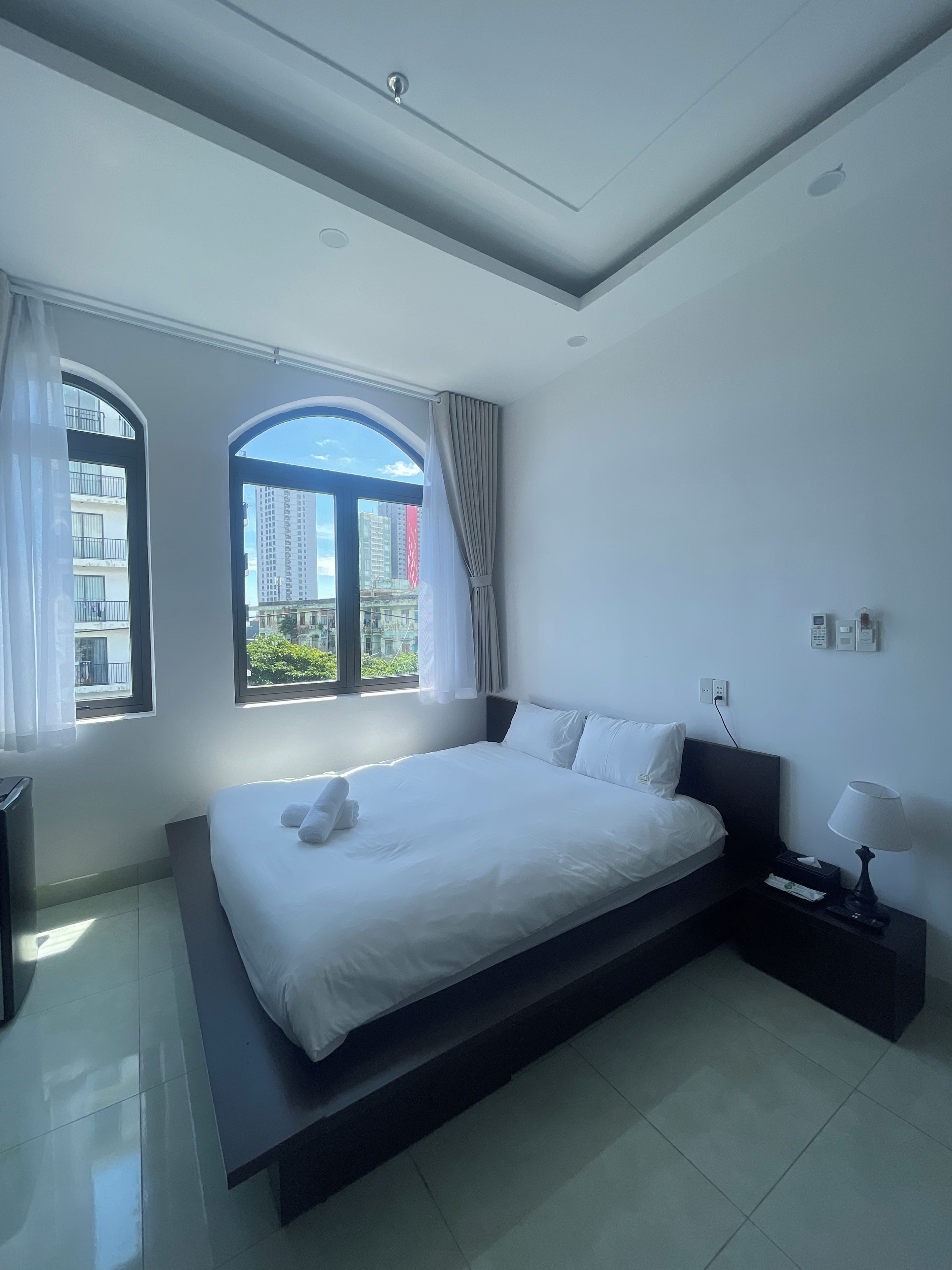 A modern hotel room with a large bed, a bedside table with a lamp, and a view of the city through large windows.