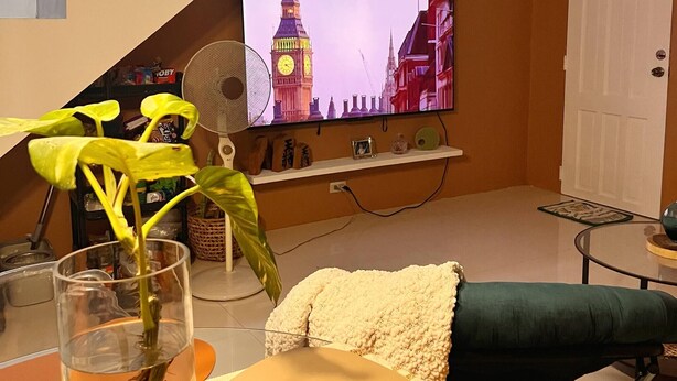 Ein Wohnzimmer mit Fernseher, der eine Stadtansicht zeigt, einer Treppe, einem Ventilator, einer Pflanze und einem Sofa.