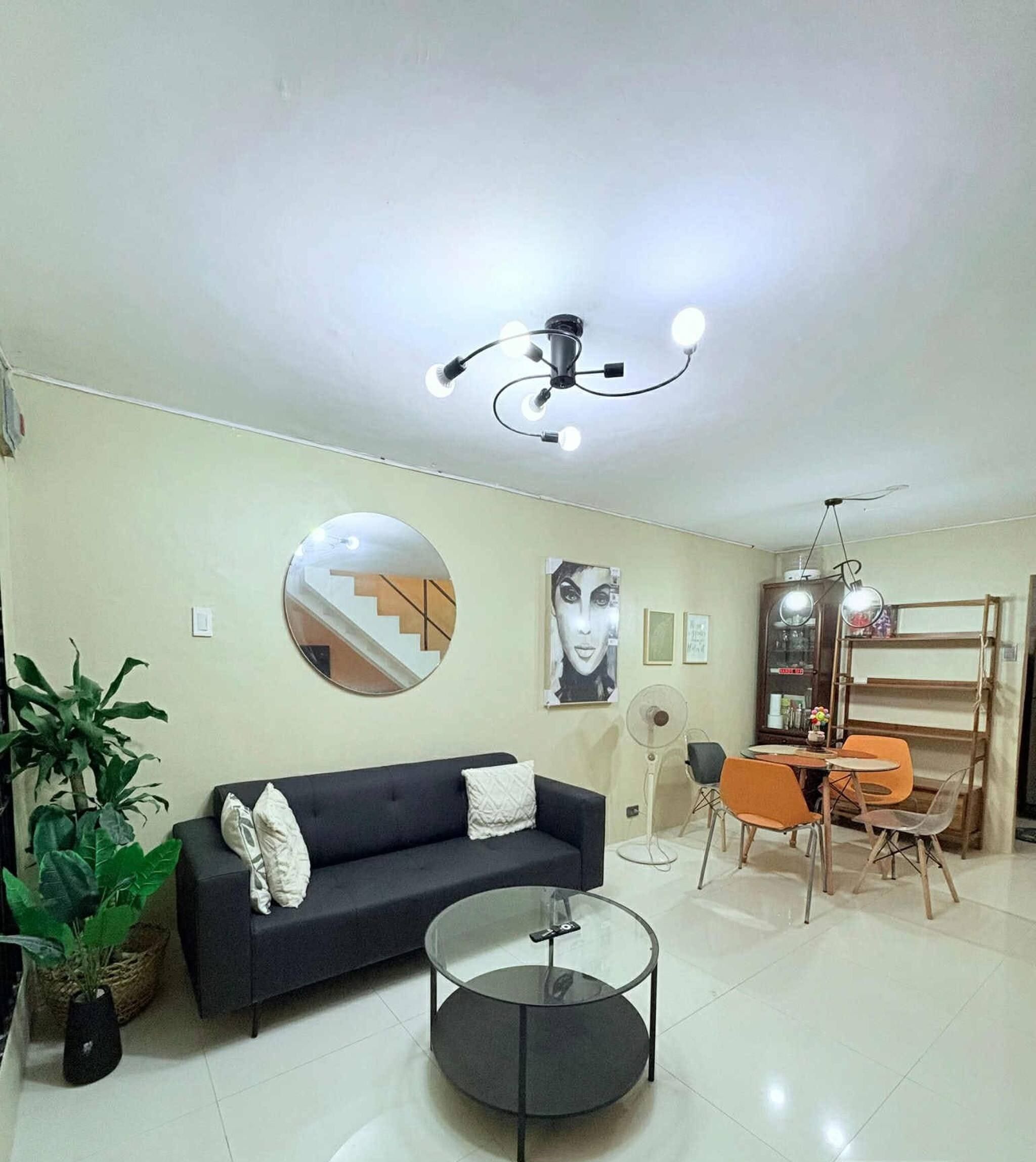 A modern living room with a sofa, a glass coffee table, a dining area with chairs, a fan, and a staircase in the background.