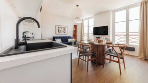 A modern kitchen with a black sink, a dining area with wooden chairs, a TV on the wall, and large windows. - Luminous Apartment - Saint Germain des Pres (Paris)