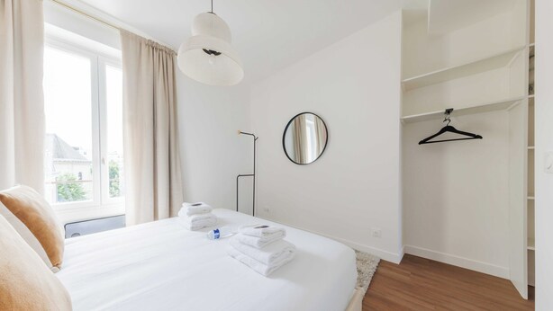 A hotel room with a large bed, a bedside table, a mirror, a hanging light fixture, a closet with a hanger, and a window with curtains. - Luminous Apartment - Saint Germain des Pres (Paris)