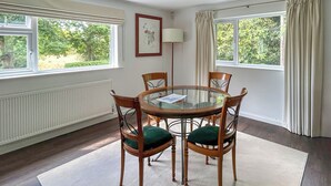 A dining area with a round glass table, six wooden chairs with green cushions, a white radiator, and two windows with curtains. - 1 bedroom accommodation in Horsham, near Brighton (Horsham, near Brighton)