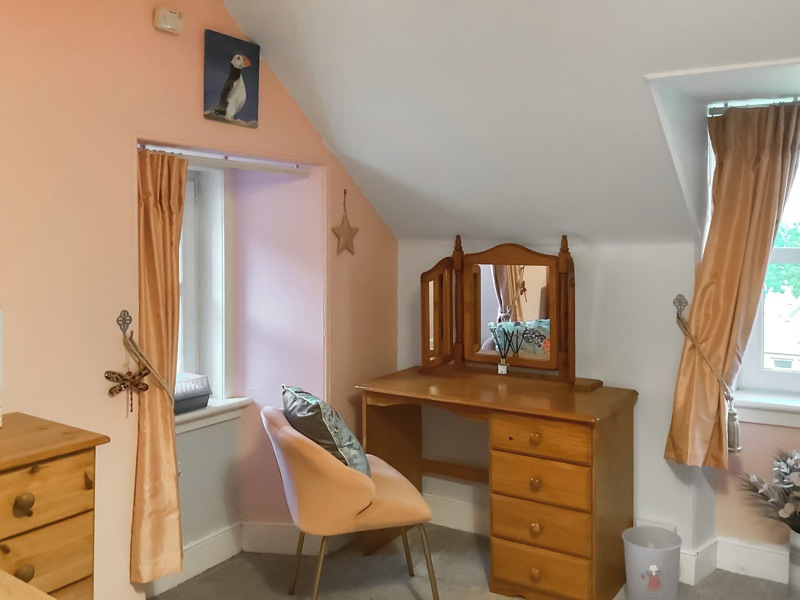 A bedroom with a sloped ceiling, a wooden desk with drawers, a chair, a window with curtains, a wall-mounted star decoration, and a pufferfish painting.