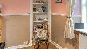 Une pièce avec une bibliothèque, un fauteuil avec un coussin, une fenêtre avec des rideaux et un radiateur.