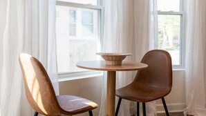A room with a round wooden table and two chairs, one with a high back and the other with a curved back, positioned near a window with white curtains.
