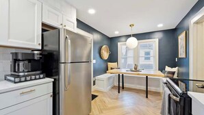 A modern kitchen with white cabinets, a stainless steel refrigerator, a coffee maker, and a dining area with a wooden table and chairs. - Close to OSU Rooftop Patio  Grill & Outdoor TV (Columbus)