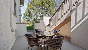 Un patio con una mesa redonda y sillas de mimbre, piso de concreto y pared blanca con una ventana.