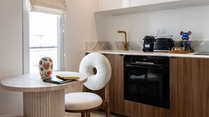 A modern kitchen with wooden cabinets, a built-in oven, and a sink with a gold faucet. There is a round table with a chair, a window with blinds, and a decorative vase on the table. - 4#New suite 4 personnes- Saint Germain Notre-Dame (Paris)