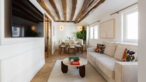 Ein modernes Wohnzimmer mit freiliegenden Holzbalken, einer Couch mit bunten Kissen, einem Essbereich mit Tisch und Stühlen sowie einem großen Fenster, das für natürliches Licht sorgt.