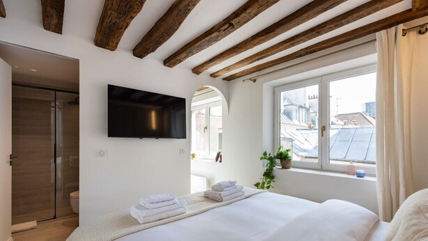 Ein Schlafzimmer mit einem großen Bett, einem an der Wand befestigten Fernseher, einem Fenster mit Blick auf Gebäude und einer Holzbalkendecke.