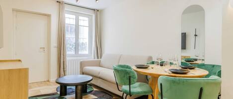 A modern living room with a round dining table, green chairs, a sofa, and a large window with curtains.