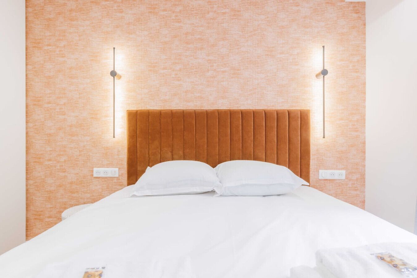 A neatly made bed with white linens and pillows, a wooden headboard, and two wall-mounted light fixtures on either side.