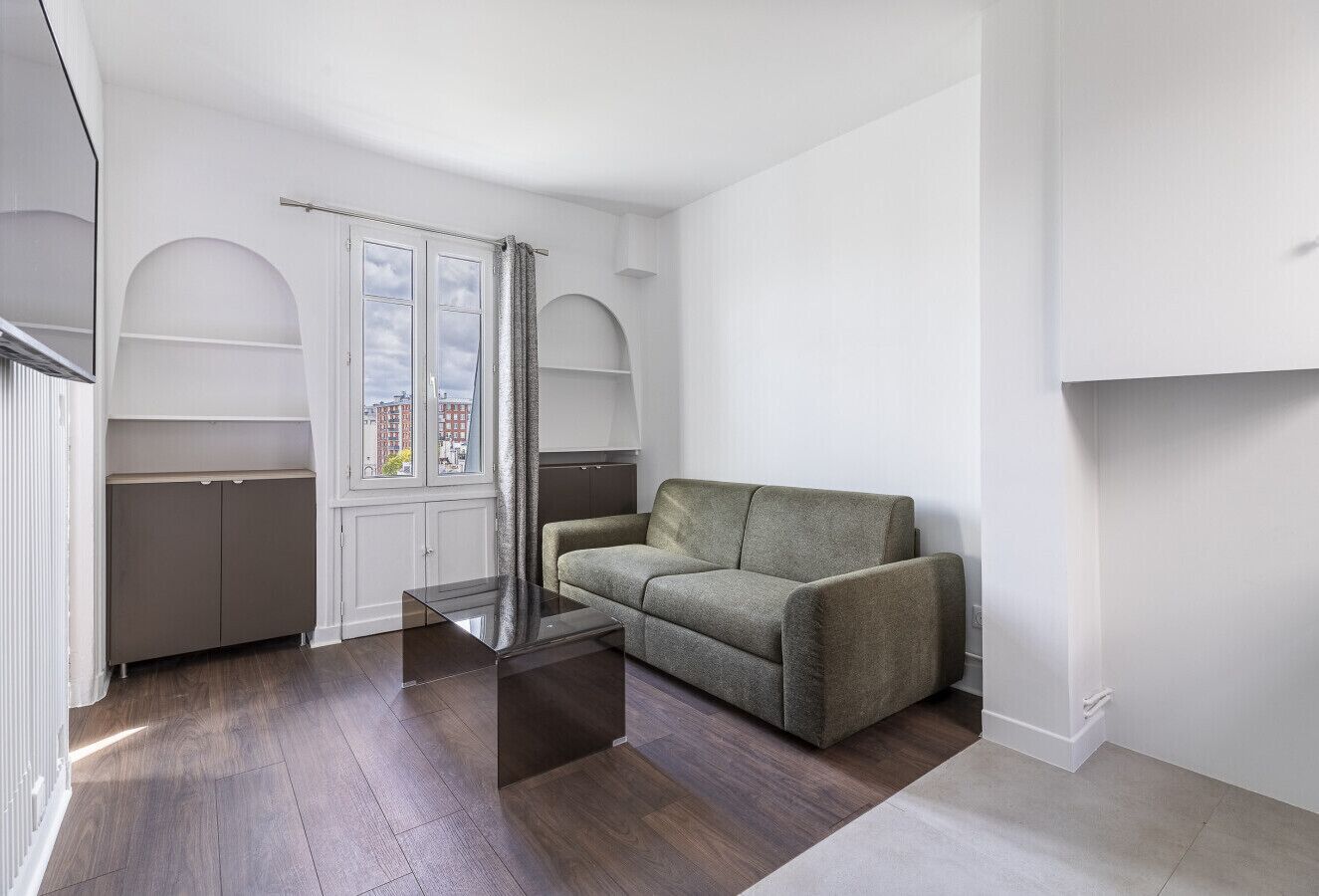 Un salon moderne avec un canapé vert, une table basse en verre et une vue sur la ville, visible à travers une fenêtre ornée de rideaux.