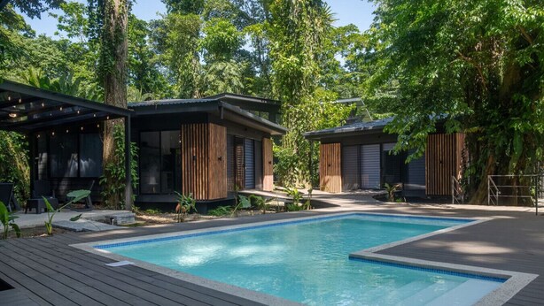A modern outdoor pool area with wooden deck flooring, two small wooden cabins, and lush greenery in the background.