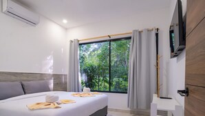 A hotel room with a bed, a TV, a window with curtains, and a view of greenery outside.
