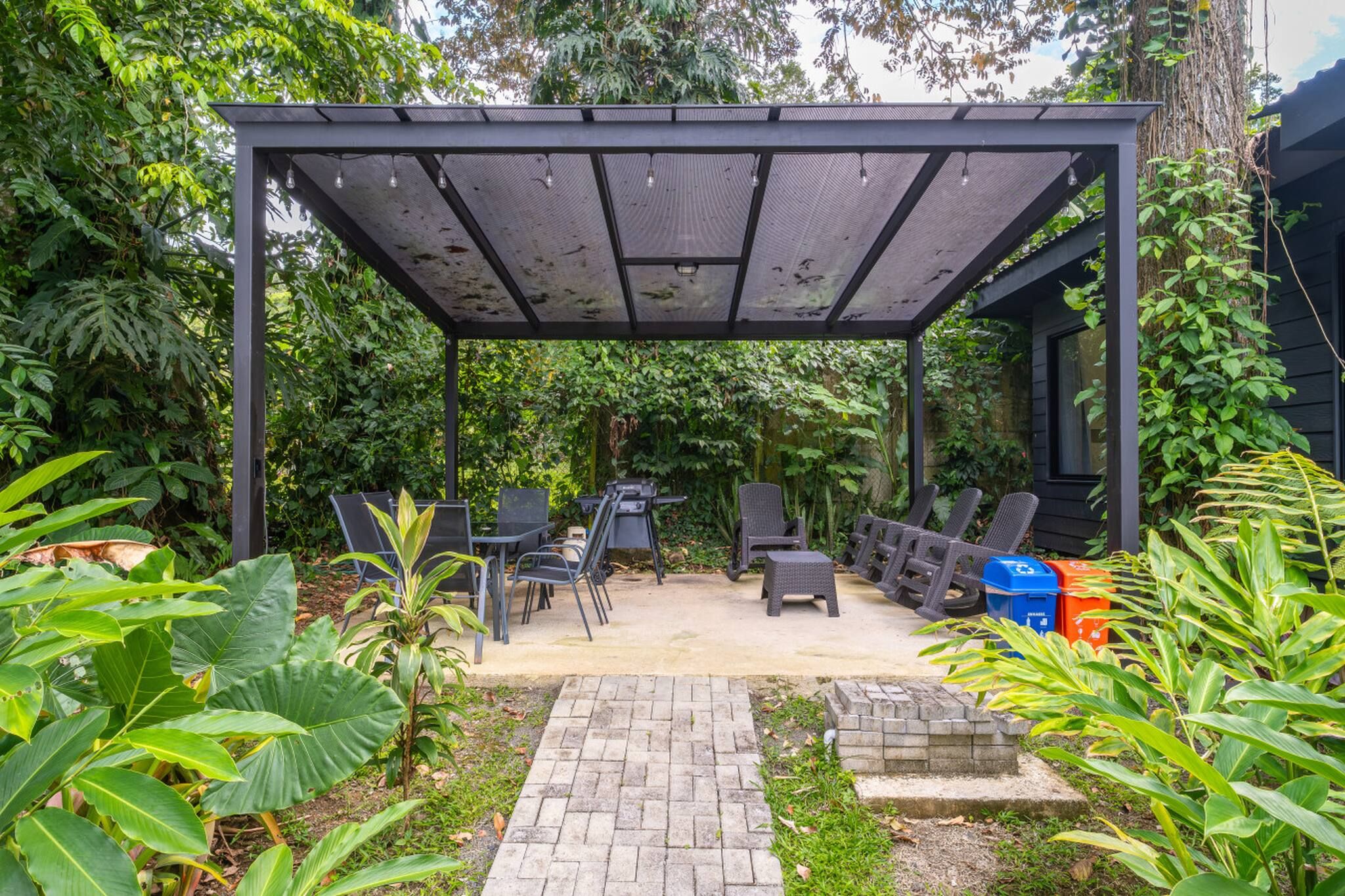 A covered patio area with outdoor furniture, a brick pathway, and lush greenery.
