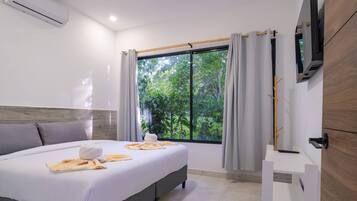 A hotel room with a bed, a TV, a window with curtains, and a view of greenery outside.