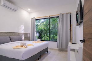 A hotel room with a bed, a TV, a window with curtains, and a view of greenery outside.