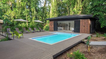 A modern outdoor pool area with a wooden deck, lounge chairs, and a small building with a kitchen visible.