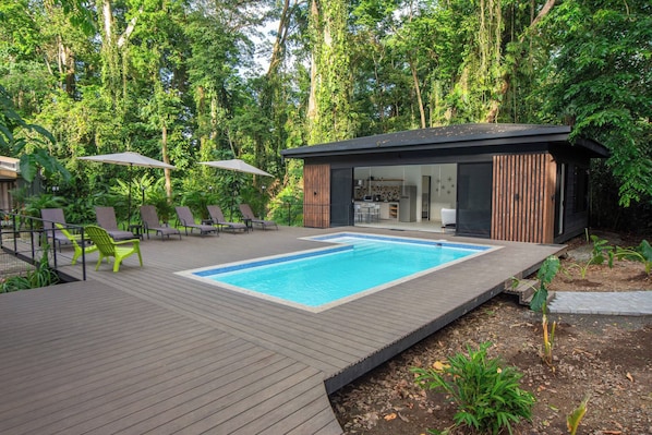 A modern outdoor pool area with a wooden deck, lounge chairs, and a small building with a kitchen visible.