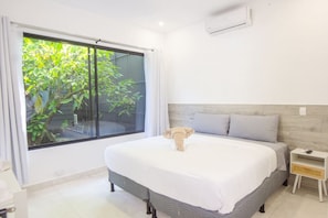 A bedroom with a large window, a bed with grey bedding, a bedside table, and a nightstand.
