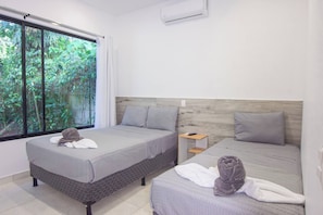A hotel room with two beds, a window with a view of greenery, and a small bedside table with a lamp.