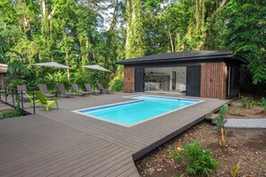 A modern outdoor pool area with a wooden deck, lounge chairs, and a small building with a kitchen visible inside.