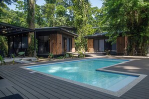 A modern outdoor pool area with a wooden deck, surrounded by lush greenery and two contemporary buildings with large windows and wooden slats.