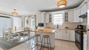A modern kitchen with white cabinets, a wooden countertop, and a built-in oven. There are two stools at the island, and a dining table with chairs is visible in the background.