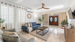 A living room with a blue sofa, a coffee table, and a ceiling fan. There is a wooden door with circular windows and a plant on the right side.