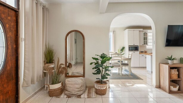 A spacious hallway with a wooden door, a large mirror, and potted plants. A dining area with a table and chairs is visible in the background.