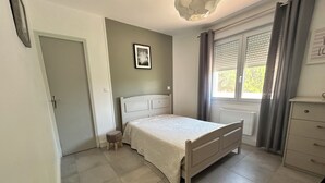 Un dormitorio con cama, un taburete pequeño, un aparador, una ventana con cortinas y un cuadro en la pared.