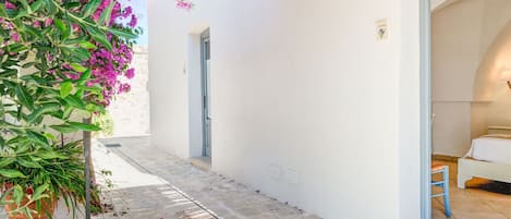 A white building with a bedroom featuring a bed, a nightstand, and a wall-mounted lamp. There is a door leading to the bedroom and a small outdoor area with a potted plant and a pathway.