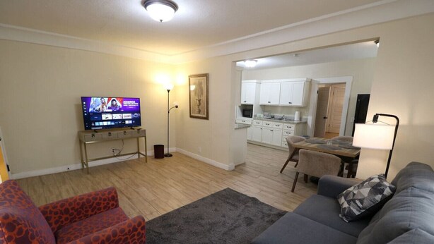 Una sala de estar con sofá, sillón y televisión sobre un mueble bajo. Se ve una cocina al fondo con armarios y microondas.