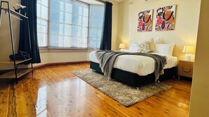 A bedroom with a wooden floor, a bed with white bedding, a ceiling fan, and a window with curtains.