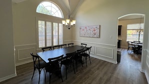 Une salle à manger avec une grande table et des chaises, un lustre et une grande fenêtre avec des stores.