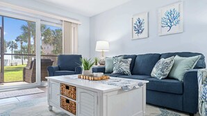 A living room with a blue sofa, a white coffee table, and a view of palm trees through a sliding glass door. - Gone Coastal l Steps to Beach l Sleeps 6 (St. Augustine)