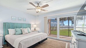 A bedroom with a bed, a ceiling fan, a nightstand, a lamp, a window with a view of a lawn and houses, and a sliding glass door. - Gone Coastal l Steps to Beach l Sleeps 6 (St. Augustine)