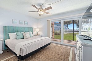 A bedroom with a bed, a ceiling fan, a nightstand, a lamp, a window with a view of a lawn and houses, and a sliding glass door.