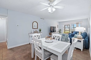 A modern living room with a dining area, a blue sofa, a TV, and a ceiling fan. - Gone Coastal l Steps to Beach l Sleeps 6 (St. Augustine)