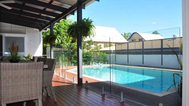 A poolside area with a wooden deck, a wicker chair, and a glass-enclosed pool.