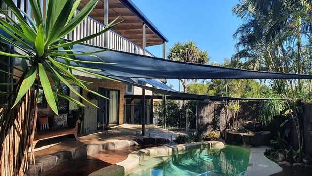 Une piscine entourée de plantes tropicales et d’une zone ombragée, avec une maison en arrière-plan.