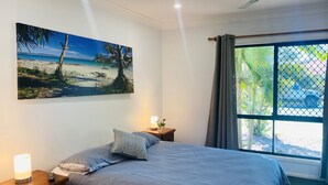 Une chambre à coucher avec un lit, une table de chevet, une lampe, un tableau représentant une plage et une fenêtre avec des rideaux.