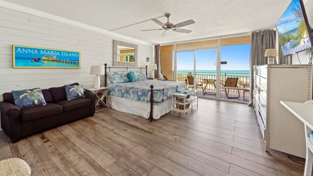 Ein Schlafzimmer mit Bett, Deckenventilator, Nachttisch, Kommode, Fernseher und einem großen Fenster mit Blick auf den Strand.