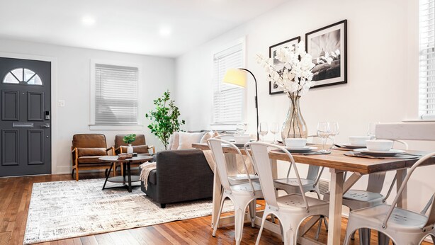 A modern living room with a dining area, featuring a wooden floor, a grey sofa, white chairs, a small table, and a vase with flowers.