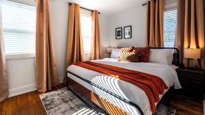 A modern bedroom with a large bed, beige curtains, a wooden floor, and a nightstand with a lamp.