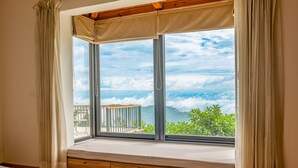 A room with a large window, a wooden bench, and a view of greenery and clouds.